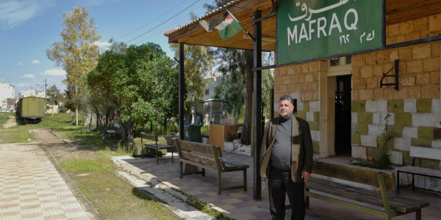 A man stands outside a train station with a sign that reads "MAFRAQ" in English and Arabic